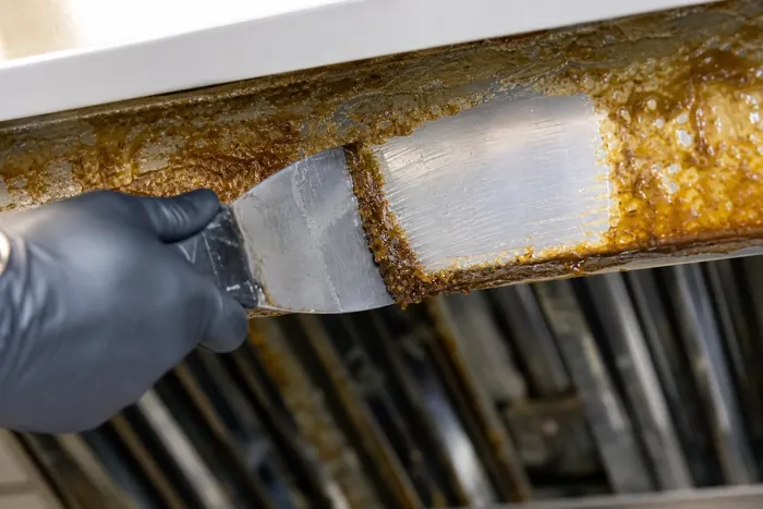 Gloved hand scraping thick baked-on grease off a duct edge before cleaning